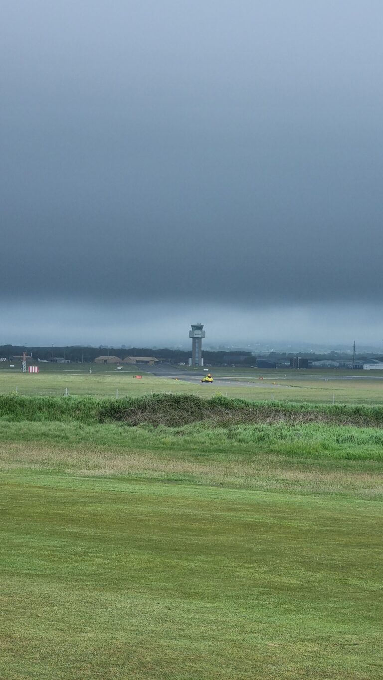 Airfield with a distant control tower and hangars beneath a low, dark storm cloud layer; green grass in the foreground.