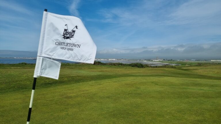 White flag on a golf green reads 'Castle Town Golf Links' with a coastal landscape in the background under a blue sky.