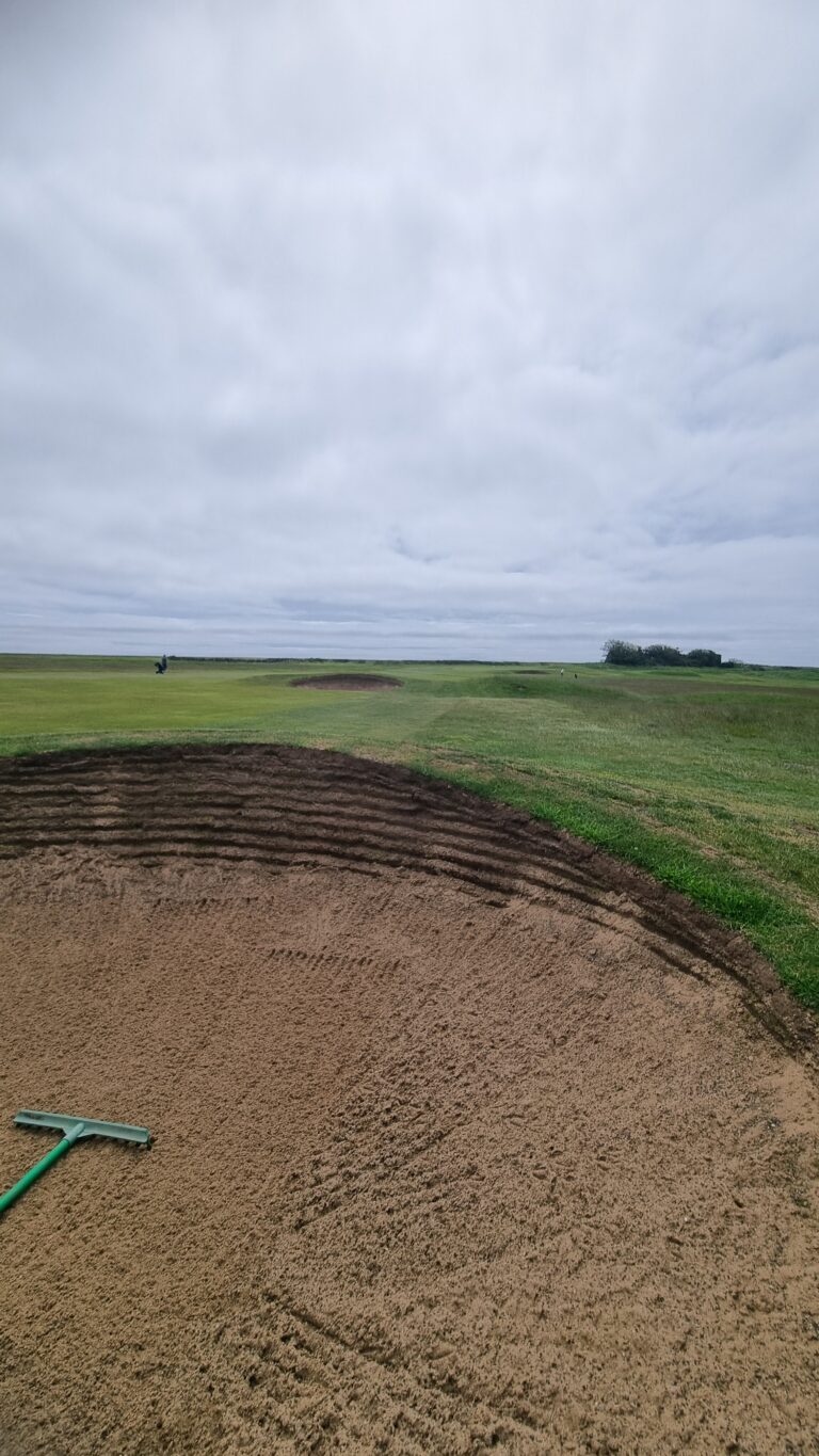 Golf course bunker in the foreground with raked sand, green fairway beyond, under a cloudy sky.