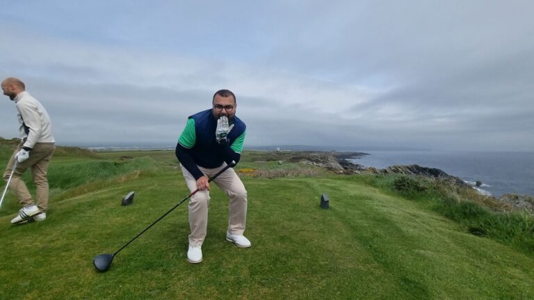 Two golfers on a coastal green; one man in a blue vest and green sleeves crouches with a glove near his mouth while holding a club, another man walks away in the background.