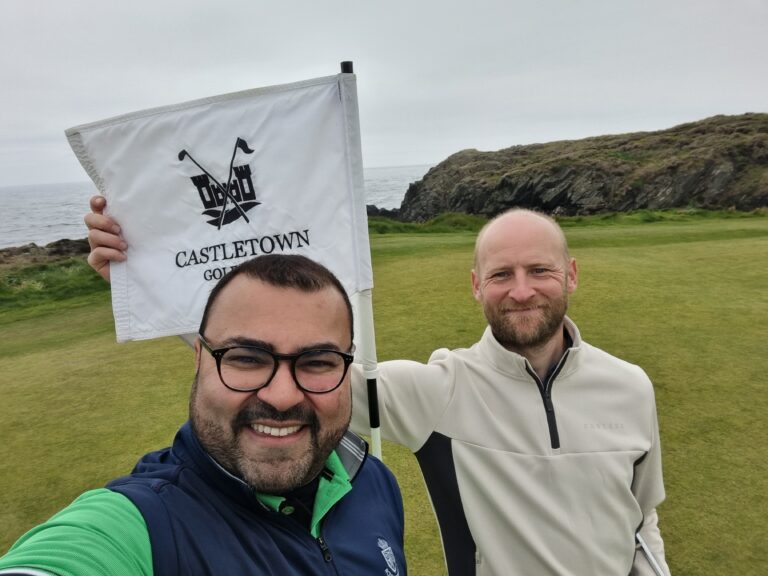 Three men take a selfie on a golf course with a white flag showing a logo and the words Castletown Golf, coastline in the background.