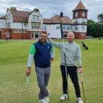 Two men pose on a golf green with clubs, smiling, flag marked '18' in view and a clubhouse behind them