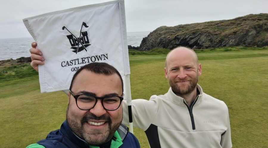Three men on a golf course taking a selfie, one holding a white flag with 'Castletown Golf' and crossed clubs logo in the foreground.