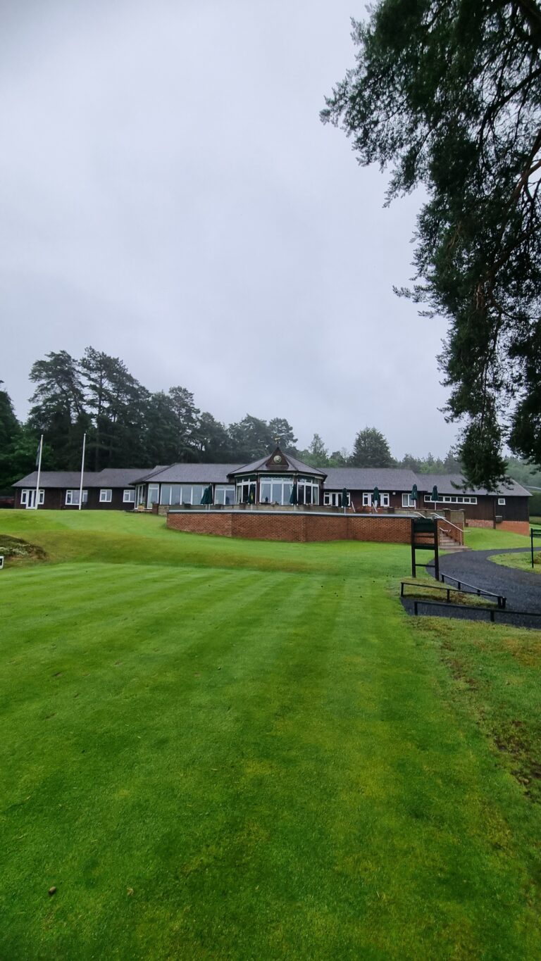Clubhouse-style building with glass-front central atrium, brick terrace, and a well-kept lawn on a cloudy day, surrounded by trees.