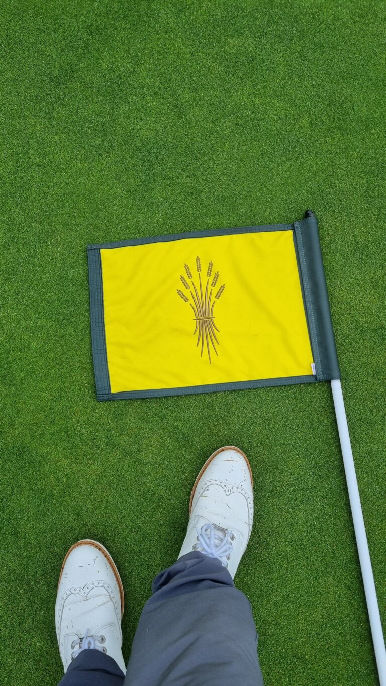Putting green scene with a yellow flag featuring a wheat stalk emblem; a white shoe is visible at the bottom of the frame.