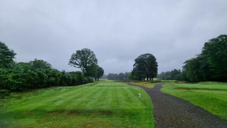 Golf course landscape with a curved gravel path, green fairways, and tall trees under a cloudy sky.
