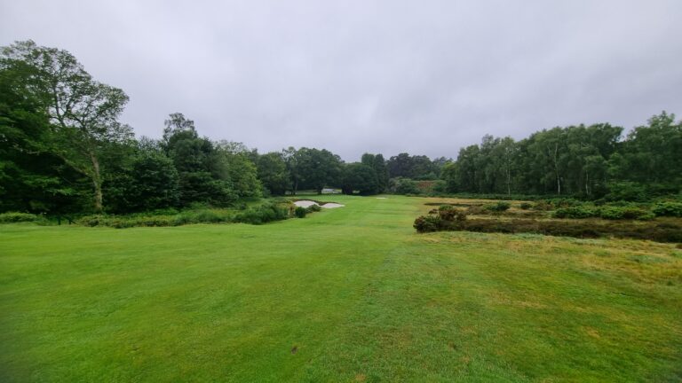 Open, grassy golf course with sand bunkers and surrounding trees under a gray, cloudy sky