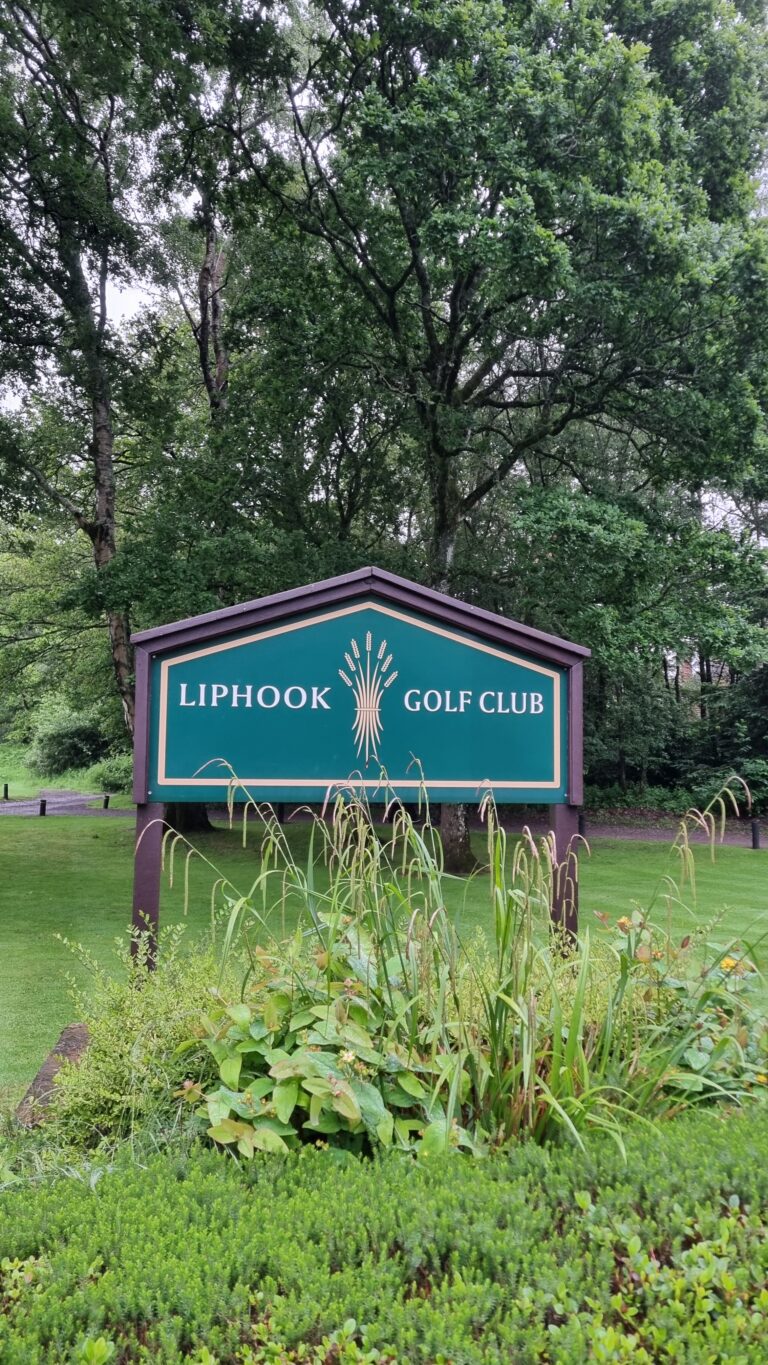 Green golf club sign reading 'Liphook Golf Club' set among trees and garden plants at the edge of a lawn.