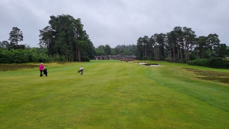 Two golfers walk across a well-kept green golf course toward a clubhouse, carrying golf bags as tall trees rise in the background.
