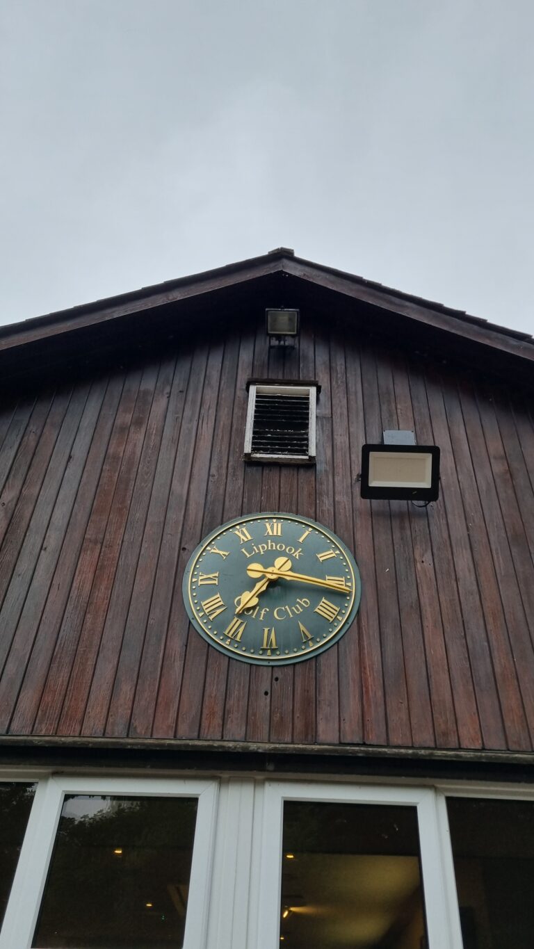 Round clock on a dark wooden clubhouse wall with Roman numerals; reads 'Liphook Golf Club' and shows about 10:10.