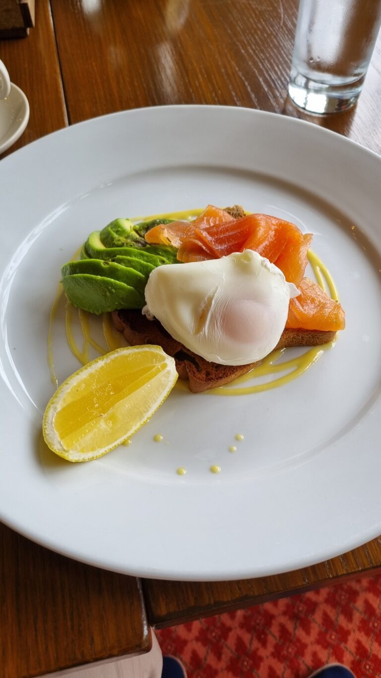 Brunch plate with avocado slices, smoked salmon, and a poached egg on toast, with a lemon wedge and dotted sauce on a white plate.