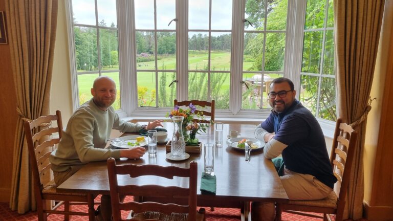 Two men sit at a wooden dining table indoors, plates and glasses in front of them, with a large window showing a green golf course outside.