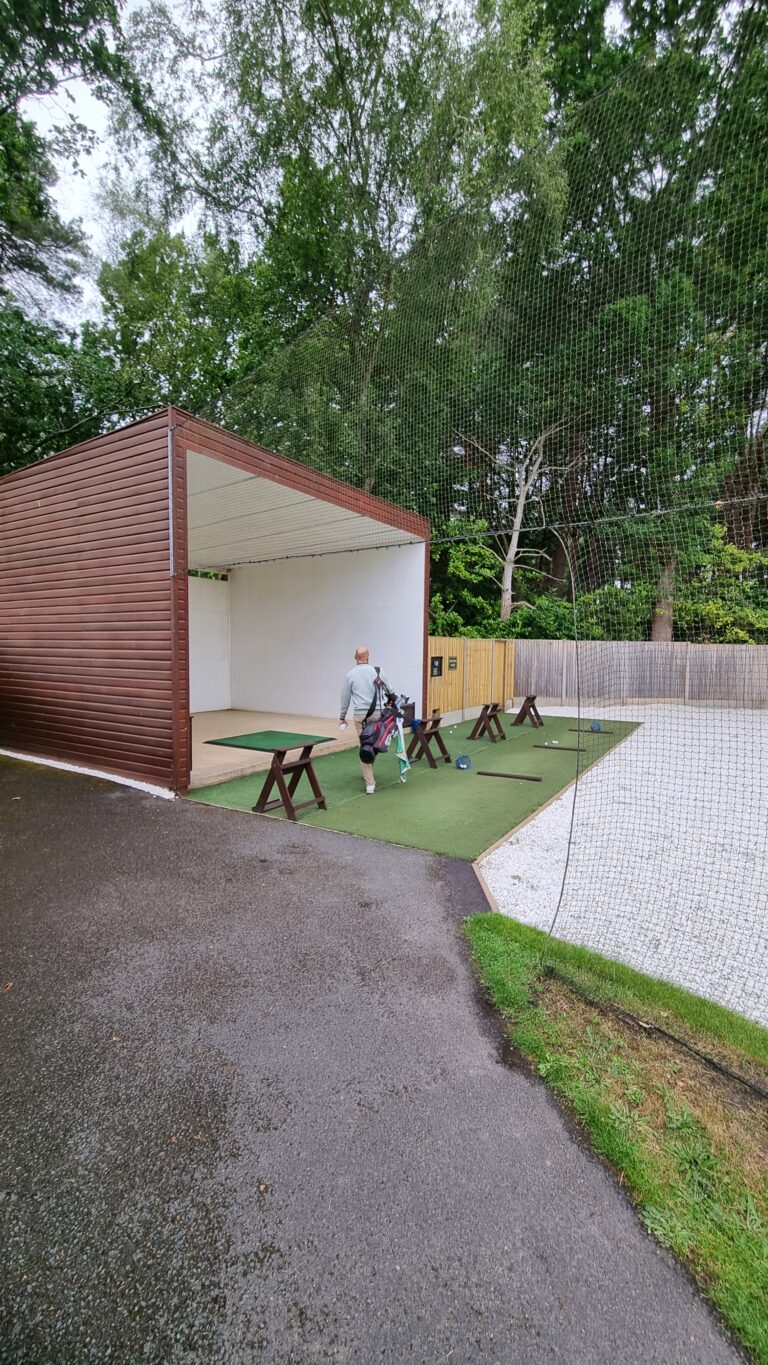 Man carrying a golf bag walks toward an open wooden shelter at a practice area with netting and benches nearby.