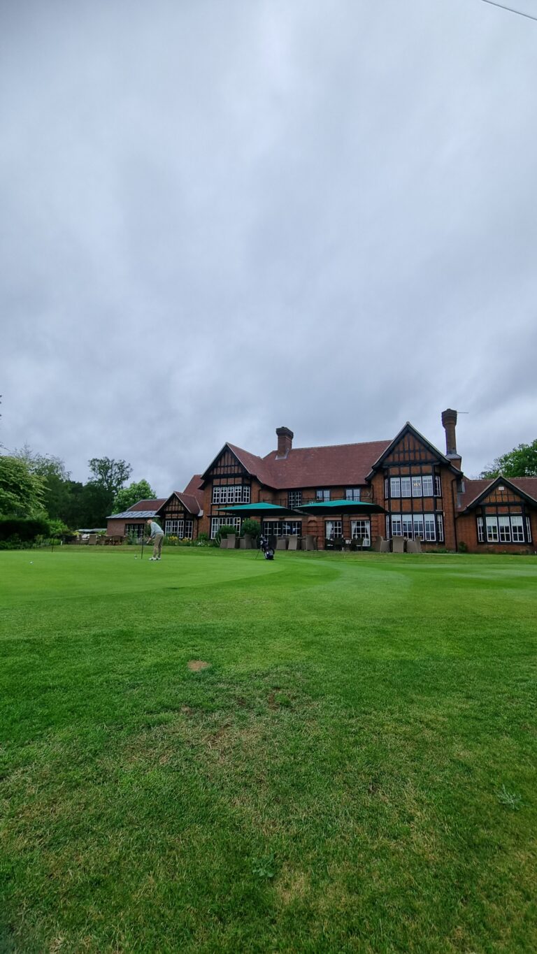 Green lawn in front of a red-brick clubhouse; a golfer is putting on the green under a cloudy sky.