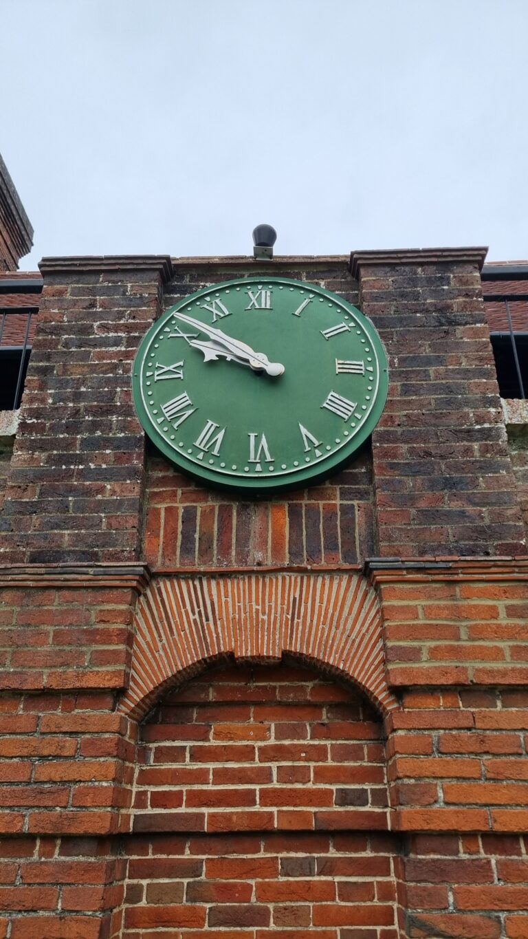 Green circular clock with white Roman numerals mounted on a brick wall facade