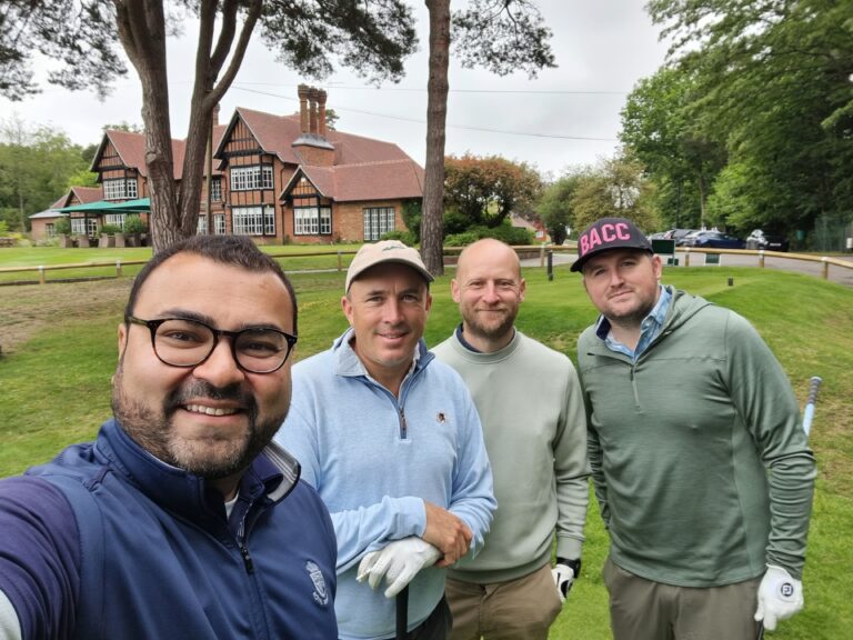 Four men pose for a selfie on a grassy lawn with a brick Tudor-style house and trees behind them.