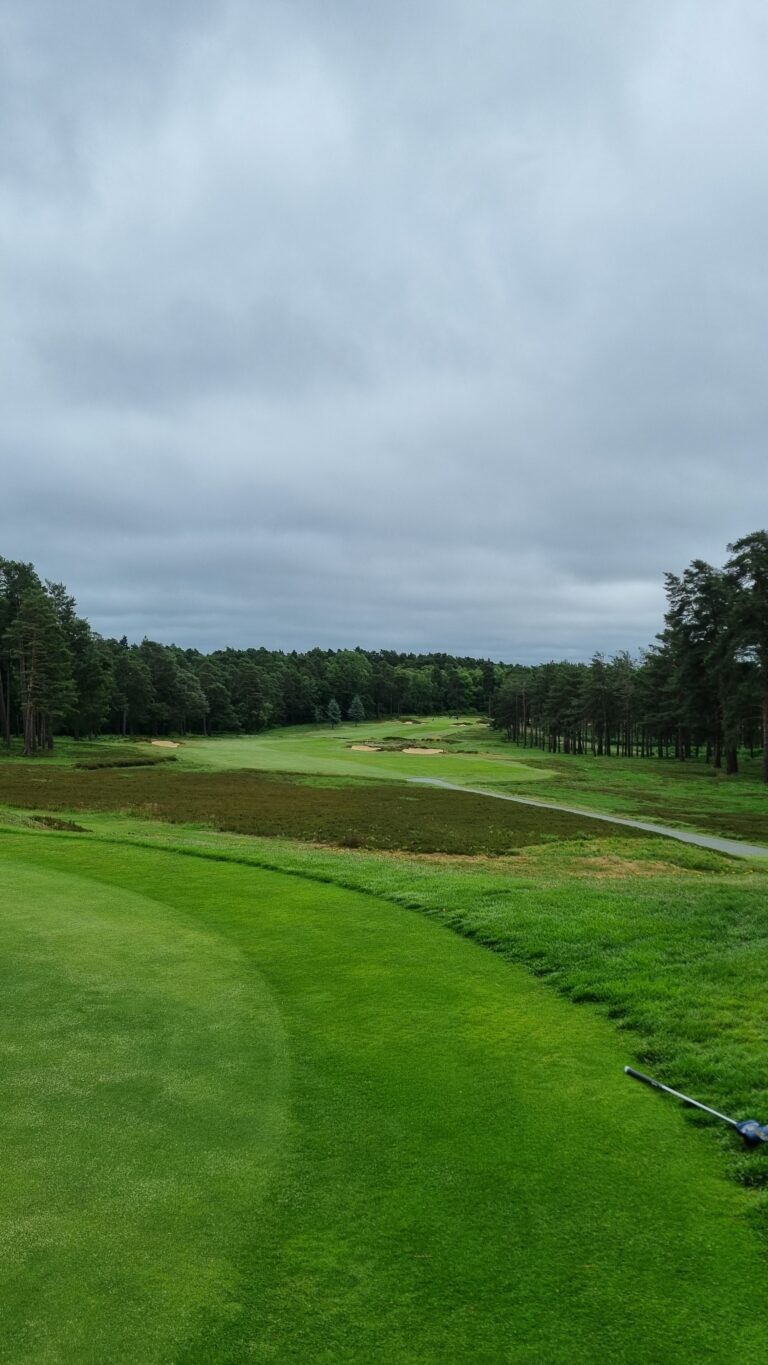 Golf course fairway lined with trees under a cloudy sky, with sand bunkers in the distance