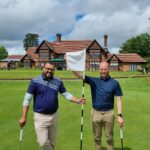 Two men standing on a golf green, holding golf clubs, with a hole and flag between them and a brick clubhouse in the background.