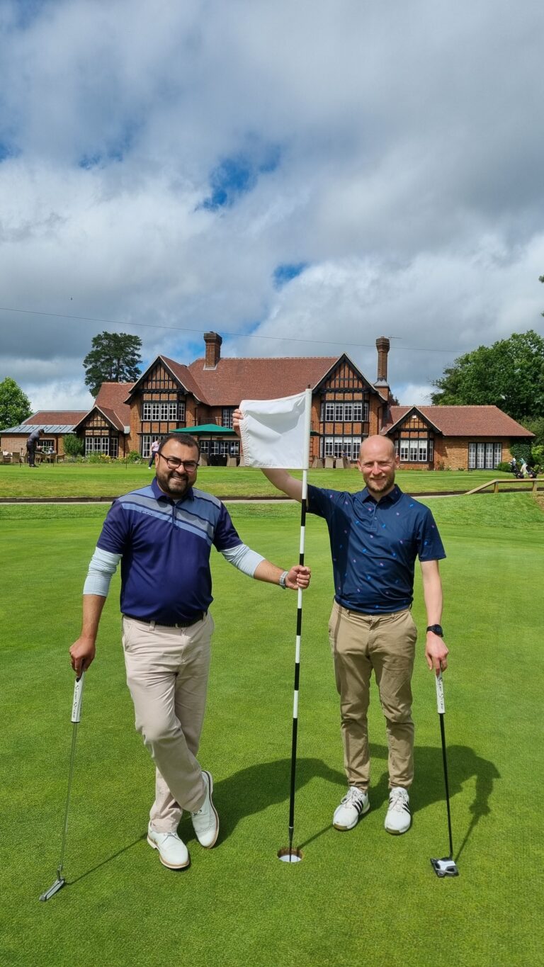 Two men standing on a golf green, holding golf clubs, with a hole and flag between them and a brick clubhouse in the background.
