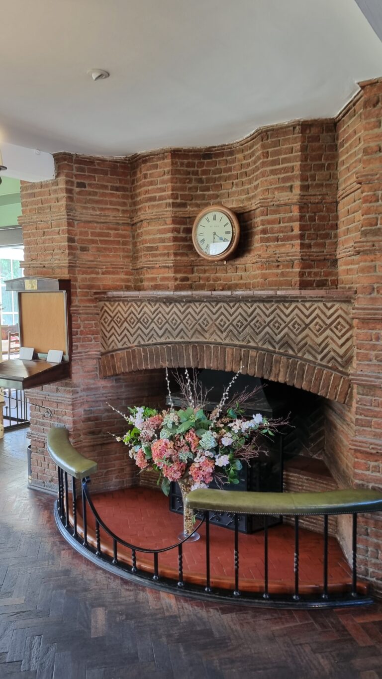 Brick fireplace with a round clock above the mantel and a bouquet of flowers in front; curved black railing in the foreground.