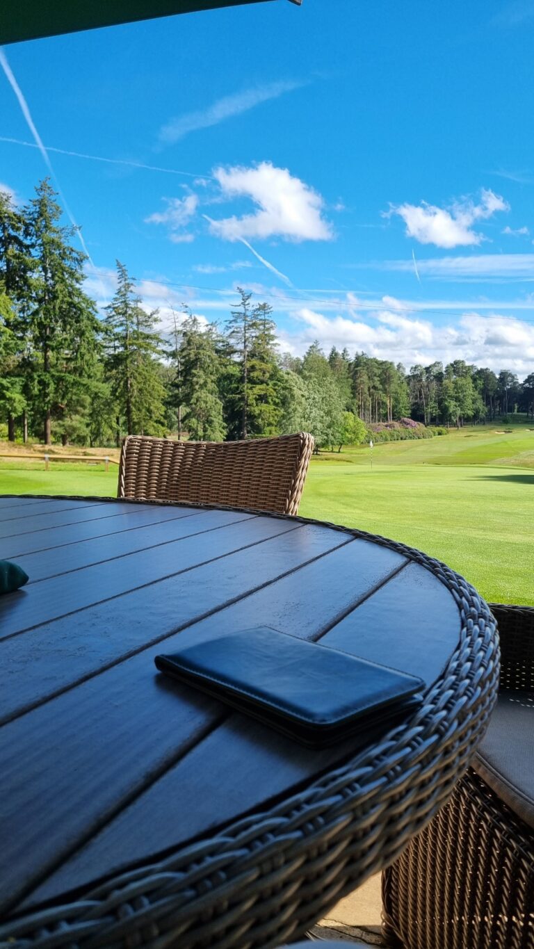 Outdoor wicker patio table with a blue surface, a closed black notebook on it, and a nearby wicker chair, with a green golf course and trees in the background under a blue sky.